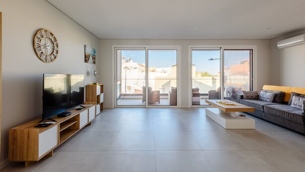 A modern living room with a flat-screen TV, wooden entertainment unit, large windows, and a grey sofa. - Alvor Ria Retreat House (Alvor)