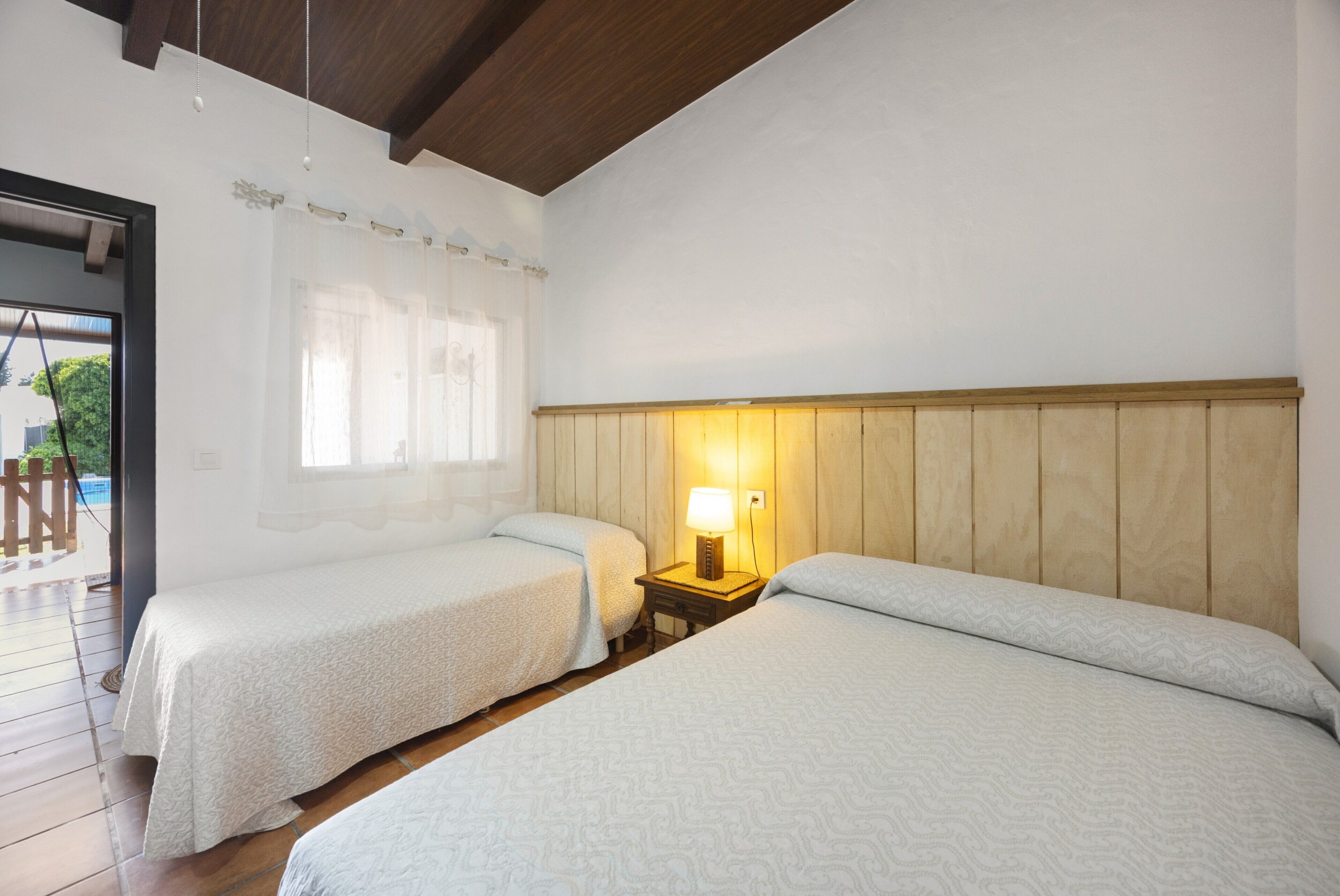 Een kamer met twee bedden, een houten hoofdbord, een nachtkastje met een lamp, een raam met gordijnen en een deur naar buiten.