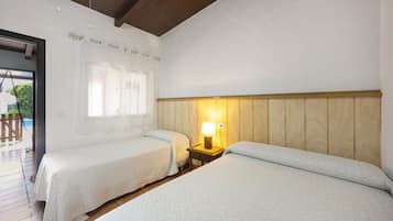 Een kamer met twee bedden, een houten hoofdbord, een nachtkastje met een lamp, een raam met gordijnen en een deur naar buiten.