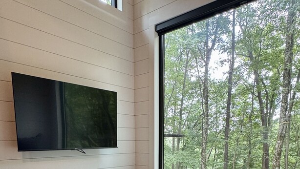 Ein Schlafzimmer mit einem Bett, einem an der Wand befestigten Fernseher und einem großen Fenster mit Blick auf den Wald.