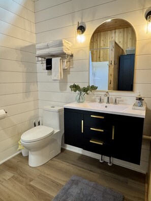 A modern bathroom with a white toilet, a vanity with black cabinets and gold handles, a mirror with a wooden frame, and a small plant on the sink.