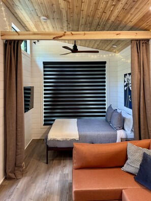A modern bedroom with a bed, a ceiling fan, a window with blinds, and a sofa.