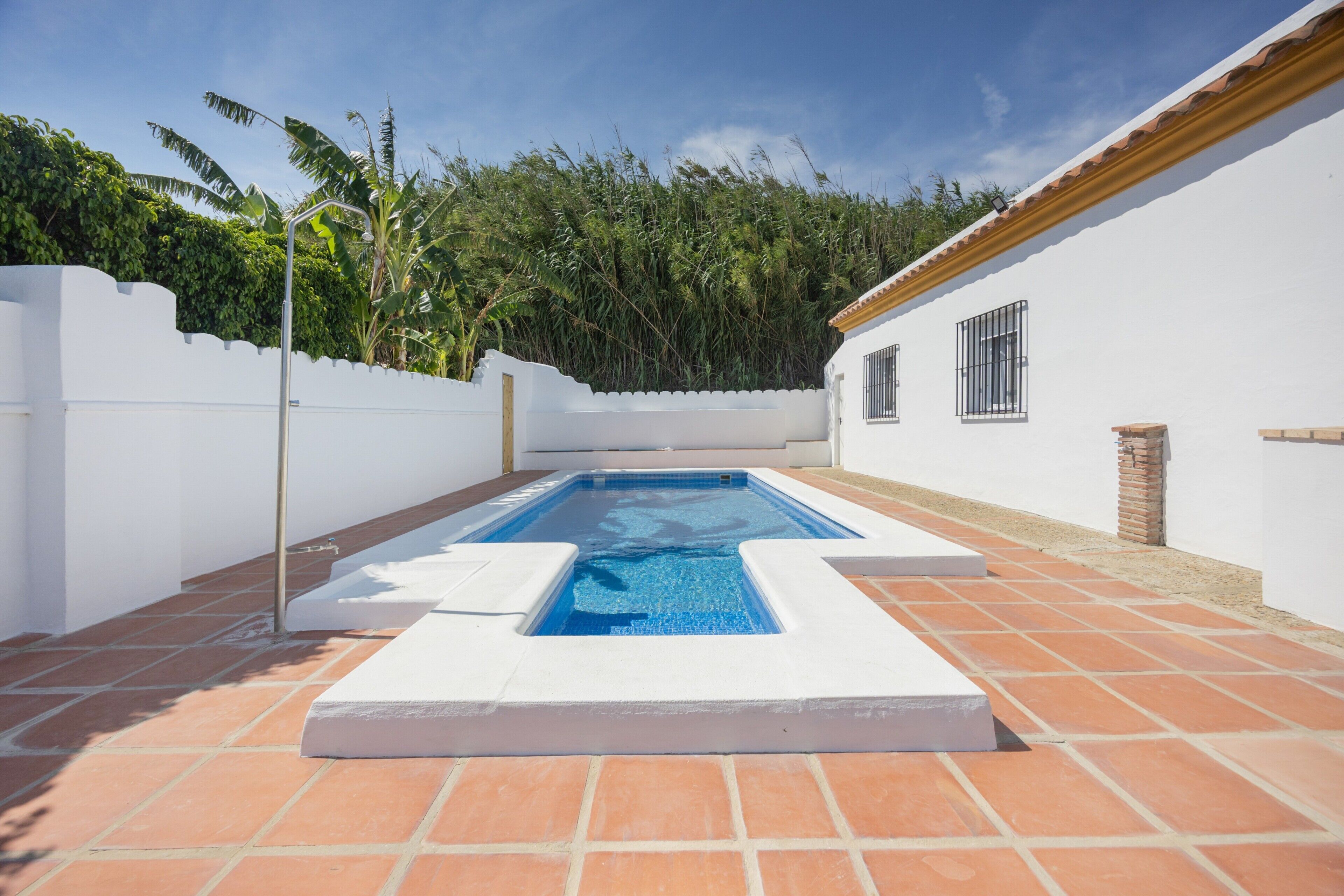 Una piscina rettangolare con bordo bianco, circondata da piastrelle in terracotta, pareti bianche e un camino in mattoni.