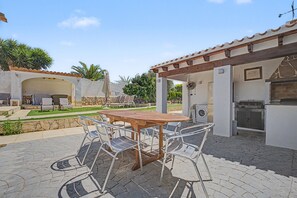 Eine Terrassengruppe im Freien mit einem runden Holztisch, Metallstühlen, einem Grill und einer Steinmauer.