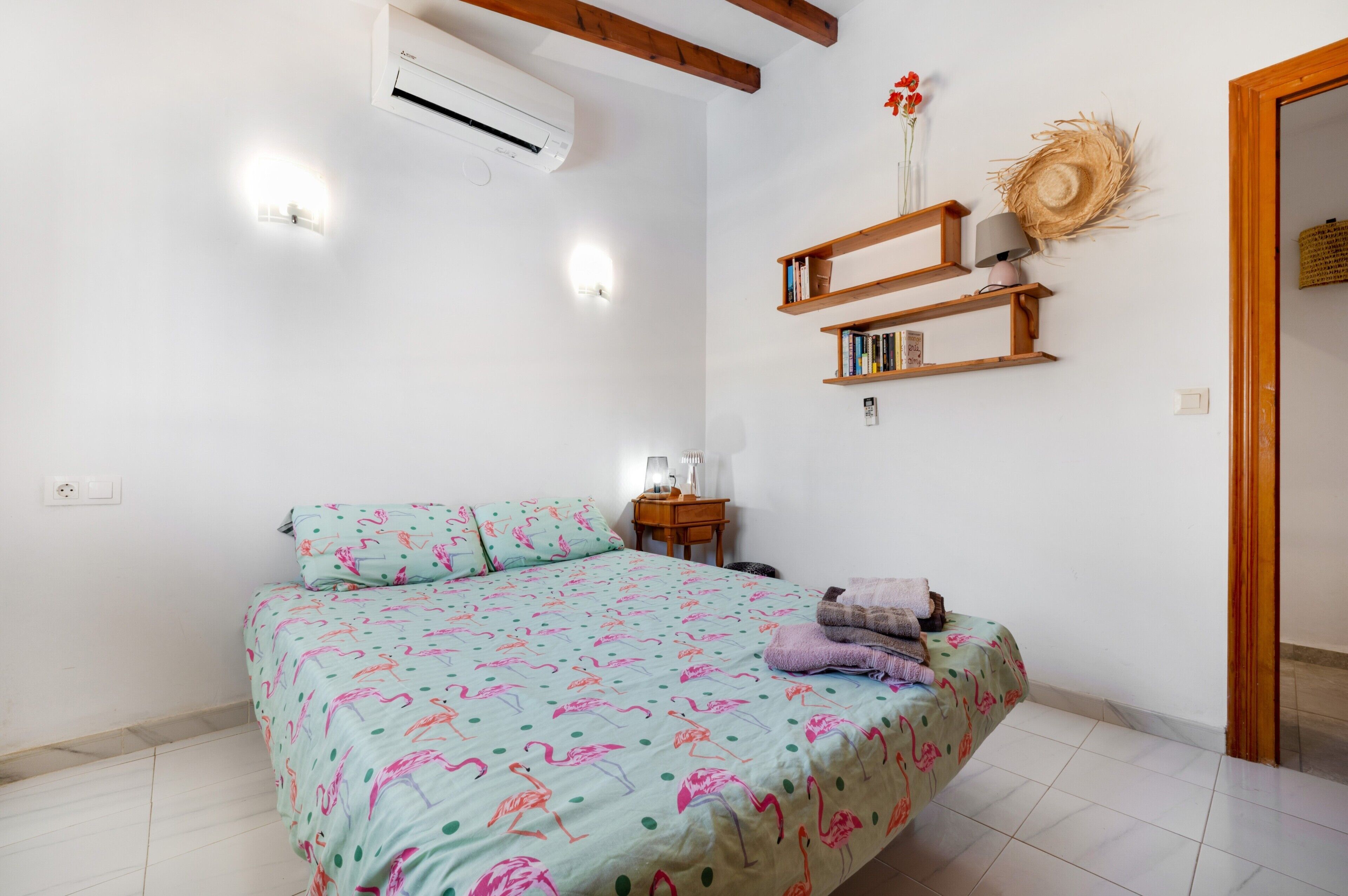 Een slaapkamer met een bed, een airconditioner, een nachtkastje met een lamp, en een wandplank met boeken en decoratieve objecten.