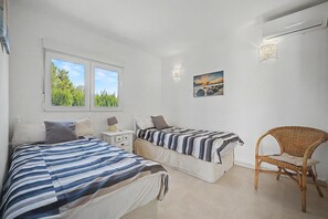 Ein Schlafzimmer mit zwei Betten, einem Nachttisch, einem Stuhl, einem Fenster mit Blick ins Grüne und einer Wandlampe.