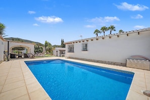 Uma piscina com um deck revestido de azulejos, uma área de estar coberta e uma parede branca com borda decorativa.