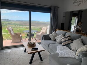 Ein Wohnzimmer mit grauem Sofa, einem kleinen Tisch und Blick auf eine grüne Landschaft durch große Fenster.