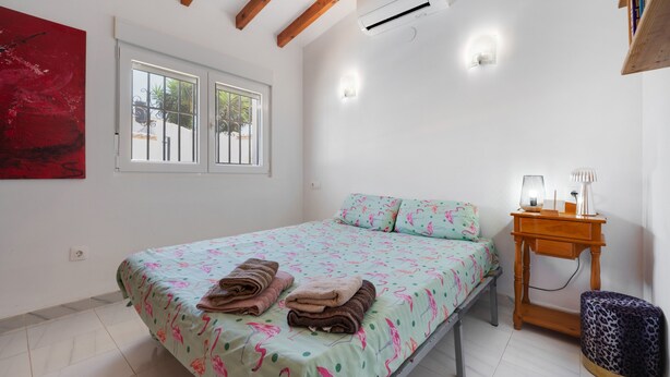 Een slaapkamer met een bed, nachtkastje met lamp, wandairconditioner en een raam met uitzicht op palmbomen.