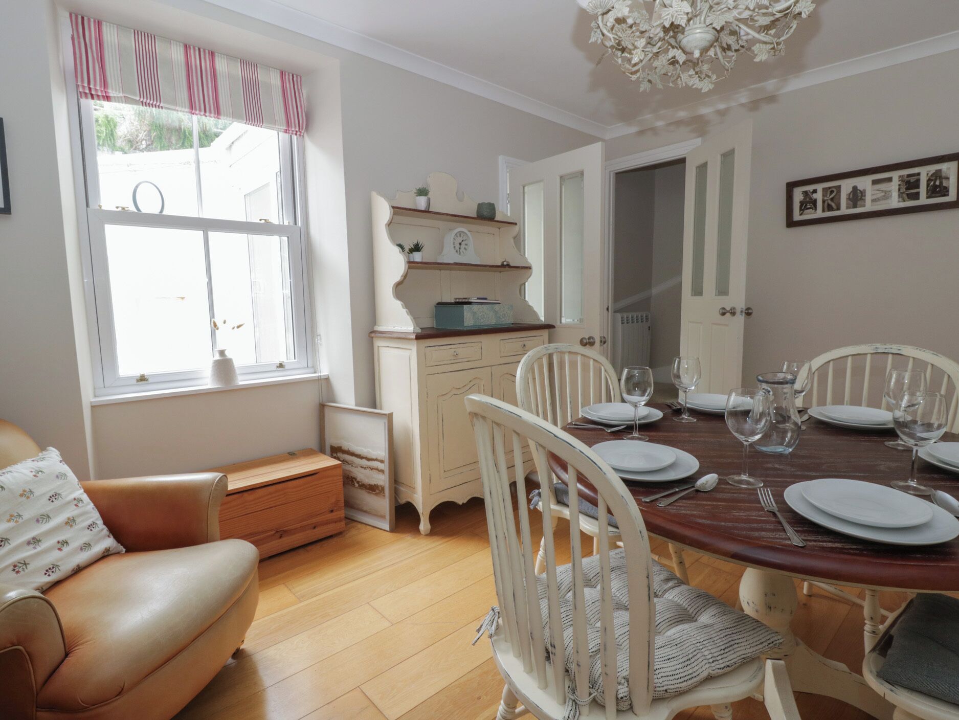 Comedor con una mesa de madera dispuesta para cuatro, sillas blancas, una ventana con cortinas, un armario blanco y una araña.