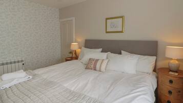 A neatly made bed with white linens, a wooden nightstand with a lamp, a radiator, and a door.