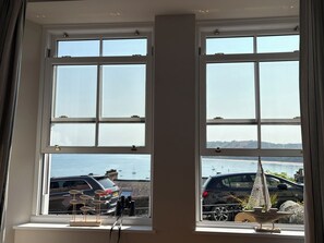 A room with a large window overlooking a body of water, a car parked outside, and a small decorative item on the windowsill. - Arosfa (New Quay)