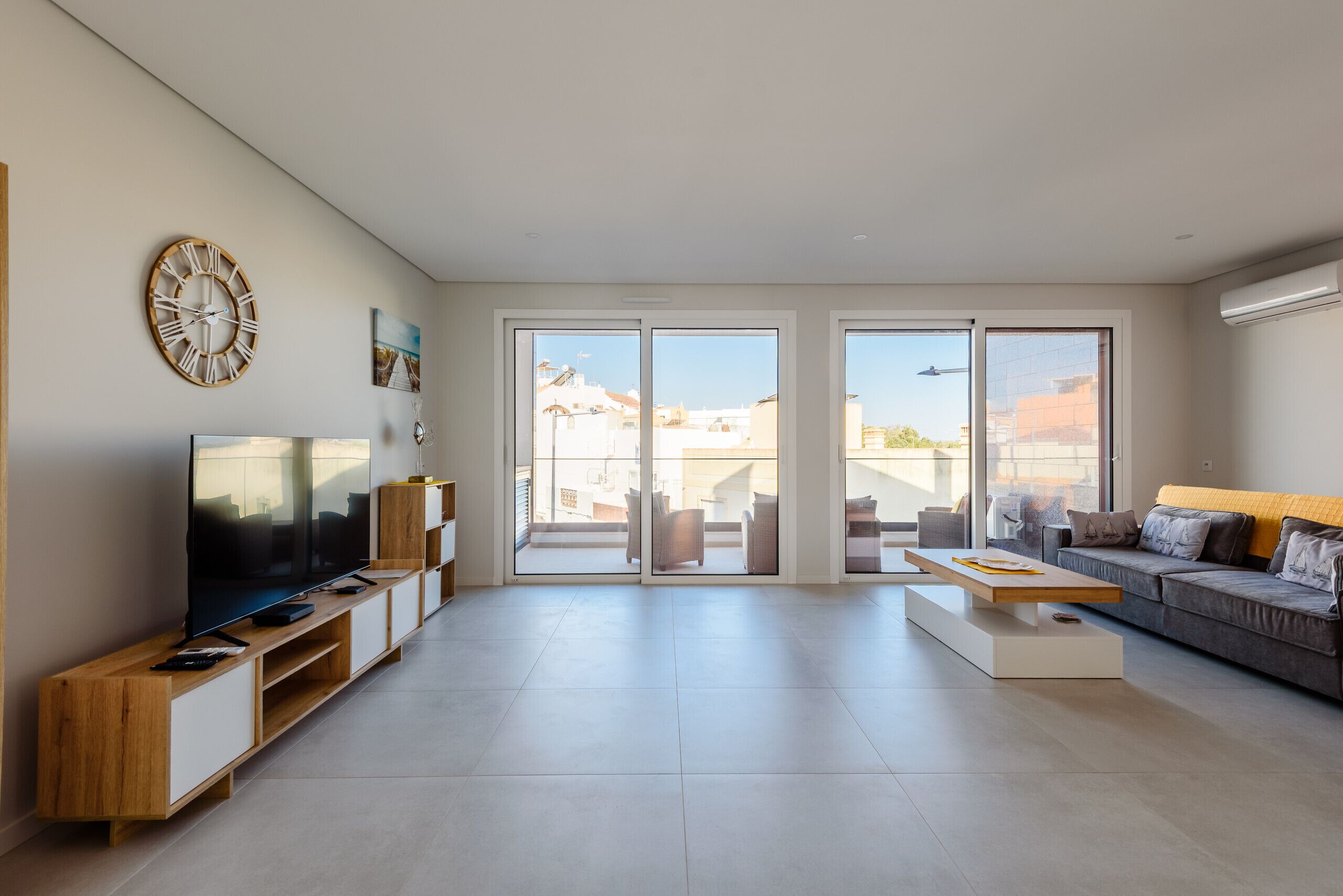 A modern living room with a flat-screen TV, wooden entertainment unit, large windows, and a grey sofa.