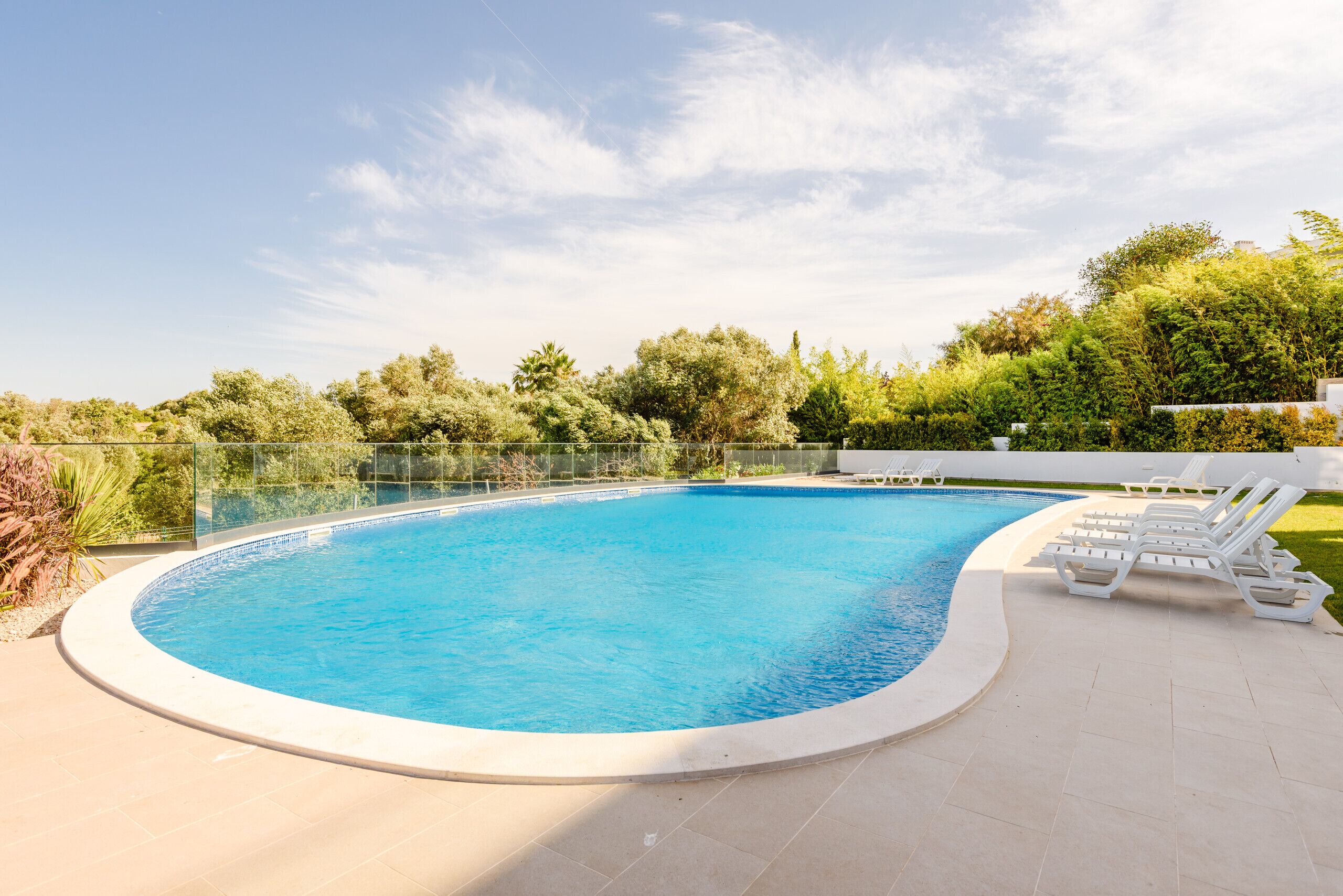 Ein Pool mit gewölbter Kante, umgeben von einer gefliesten Liegefläche und weißen Liegestühlen.