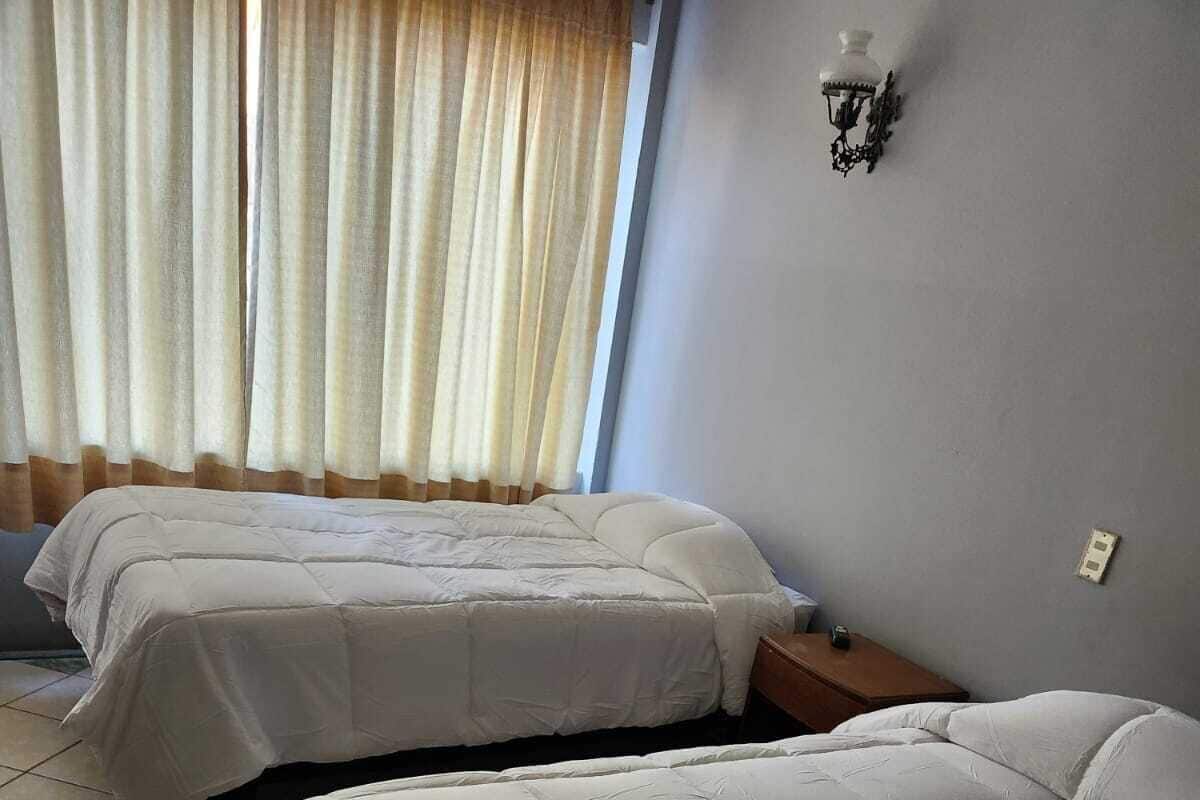 Una habitación con dos camas, una mesita de noche, una lámpara de pared y un ventanal con cortinas.