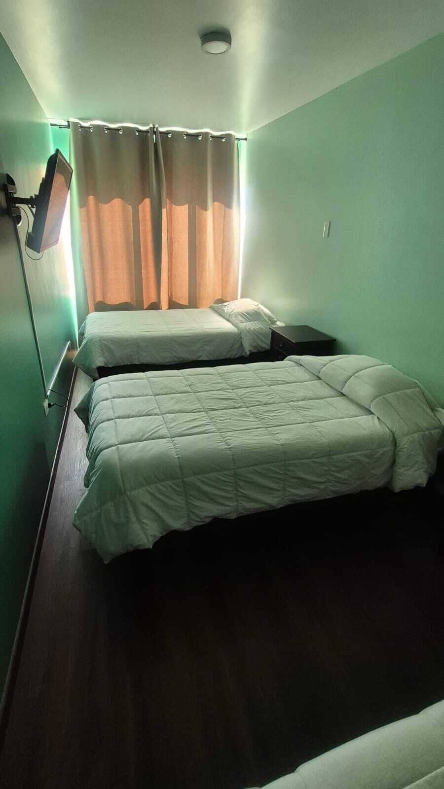 A room with two beds, a window with curtains, and a wall-mounted light fixture.
