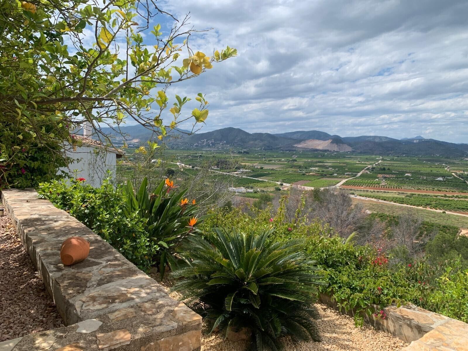 A stone wall with plants and a view of a valley with fields and mountains.