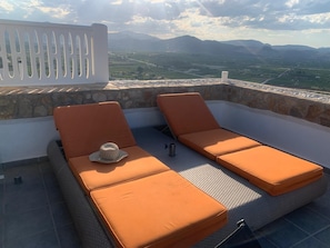 Eine Dachterrasse mit zwei orangefarbenen Liegestühlen, einem gewebten Sofa und Blick auf ferne Berge.
