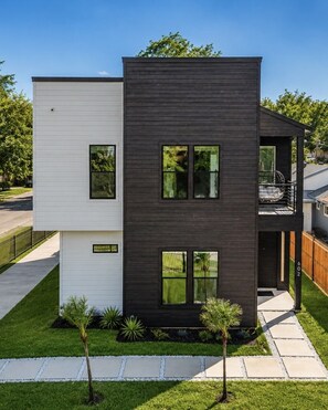 A modern two-story house with a balcony, a small garden, and a clear sky.