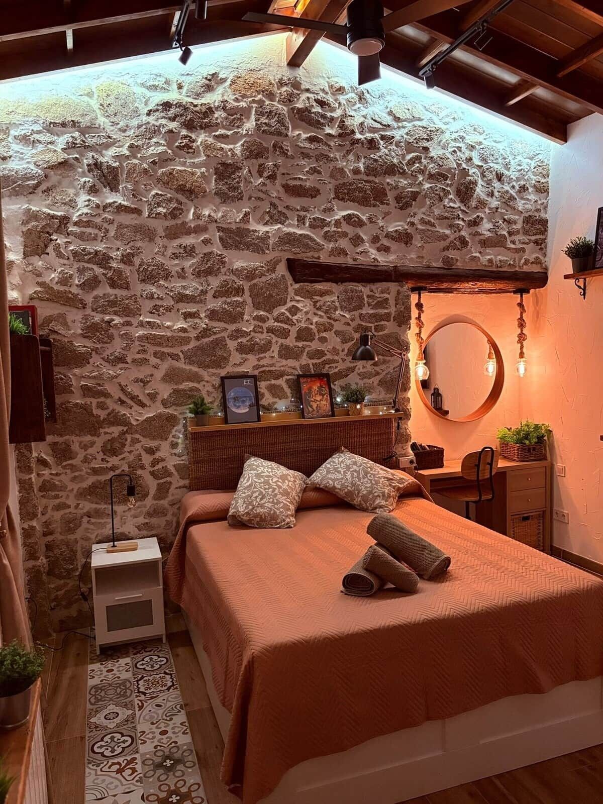 Een knus slaapkamer met een stenen muur, een bed met een decoratieve spreidek, een nachtkastje met een lamp en een klein plankje met decoratieve objecten.