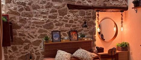 A cozy bedroom with a stone wall, a bed with a patterned bedspread, a bedside table with a lamp, and a small shelf with decorative items.