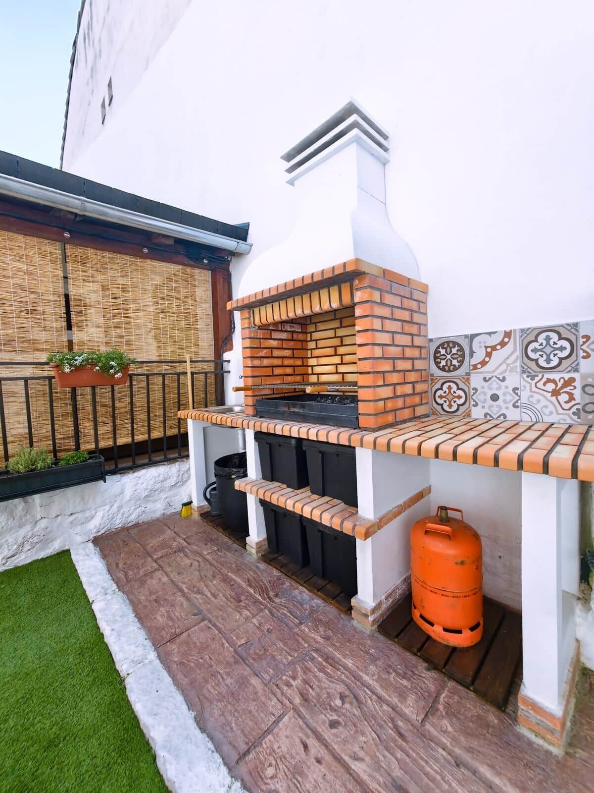 Un espace barbecue extérieur en briques, équipé d’une bouteille de gaz, d’une poubelle noire et d’un petit parterre agrémenté de plantes.