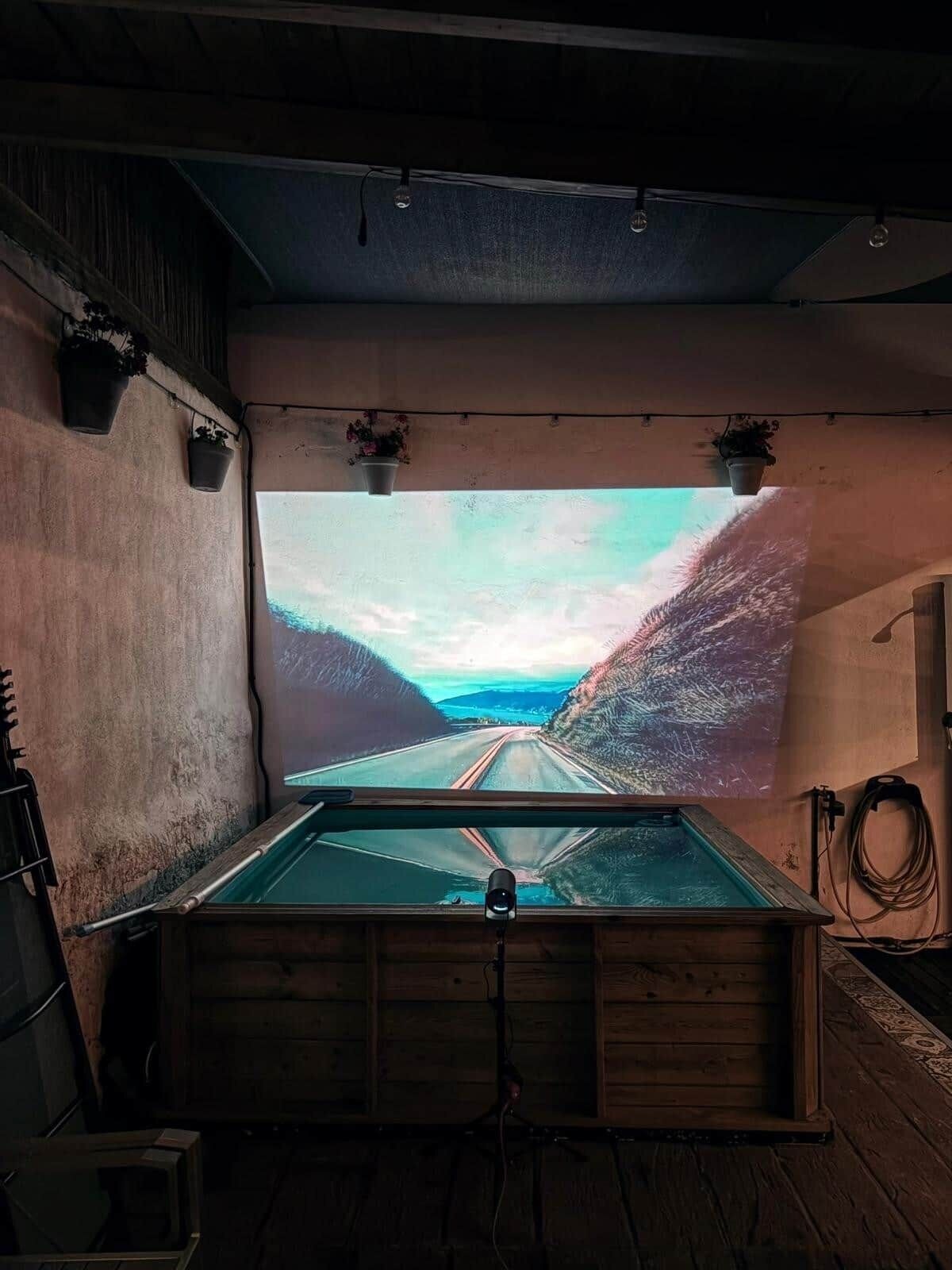 Una habitación con una gran pantalla proyectando una vista panorámica de una carretera, paredes de madera y plantas colgantes.