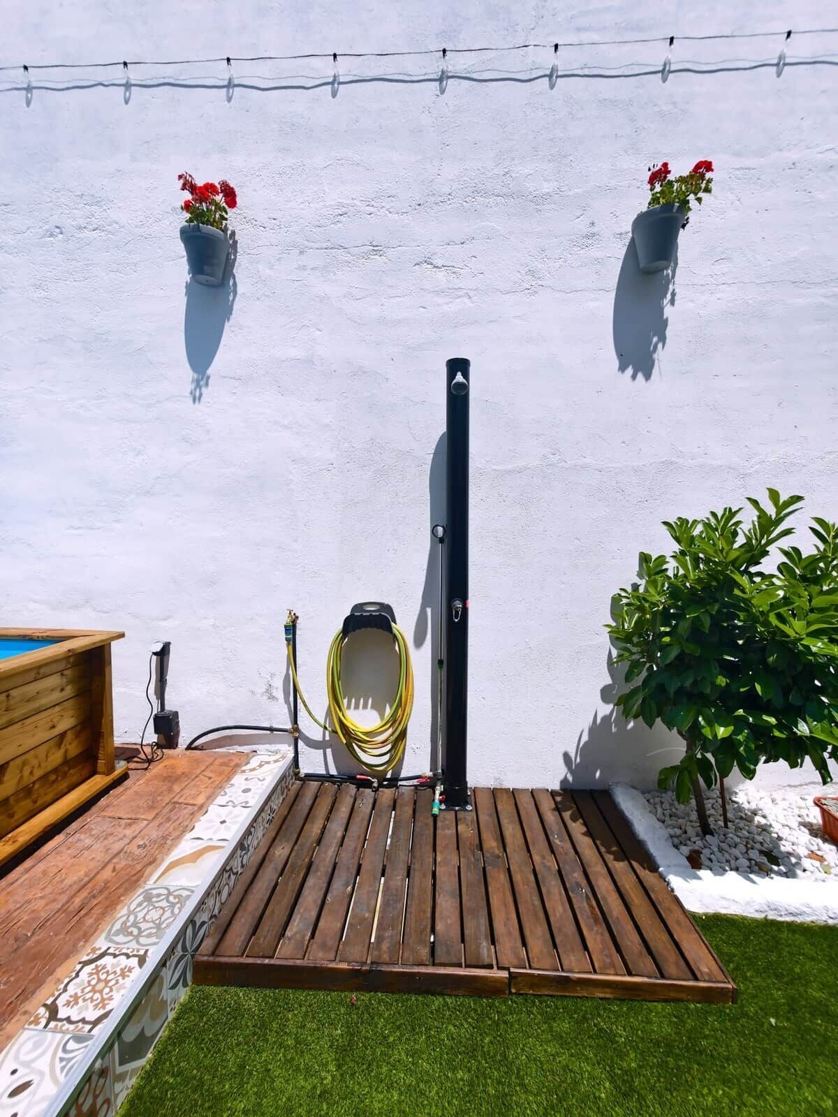 Een houten terras met een tuinslang en een zwarte paal, omgeven door een witte muur en plantenpotten.