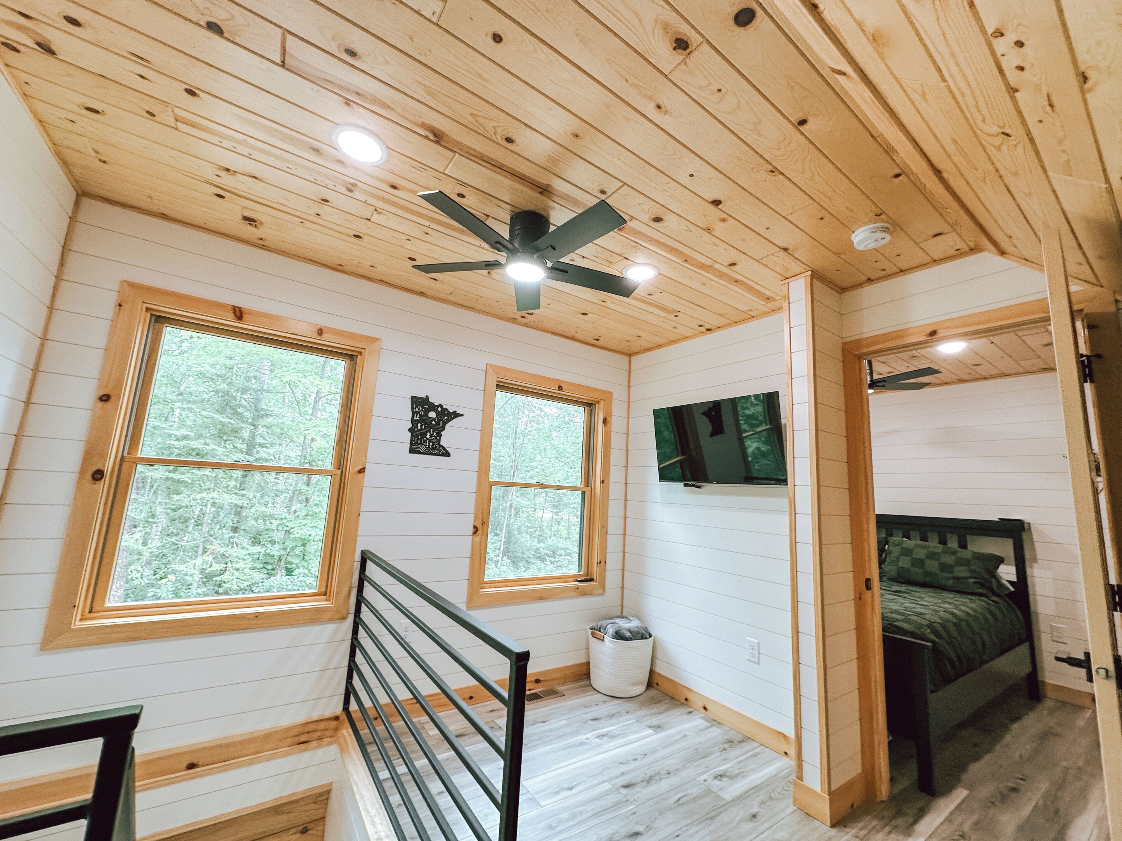 A cabin interior with wooden walls, a ceiling fan, two windows with a view of trees, a bed, and a small trash can.
