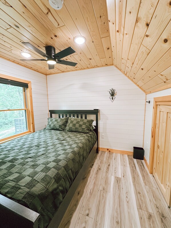 Un dormitorio con techo de madera, cama con colcha a cuadros, ventilador de techo y una ventana.