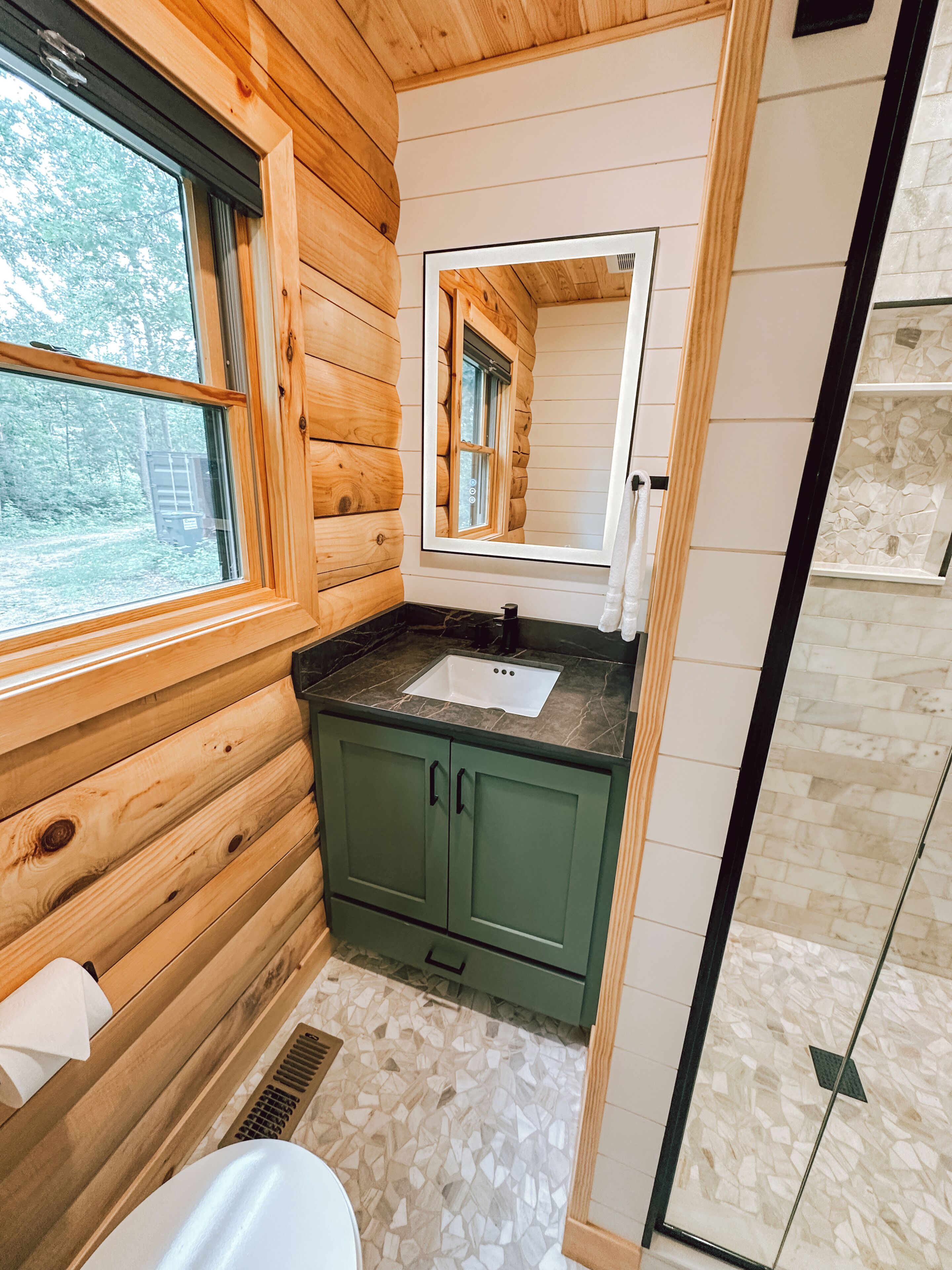 Baño de cabaña de madera con ventana, lavamanos y espejo.