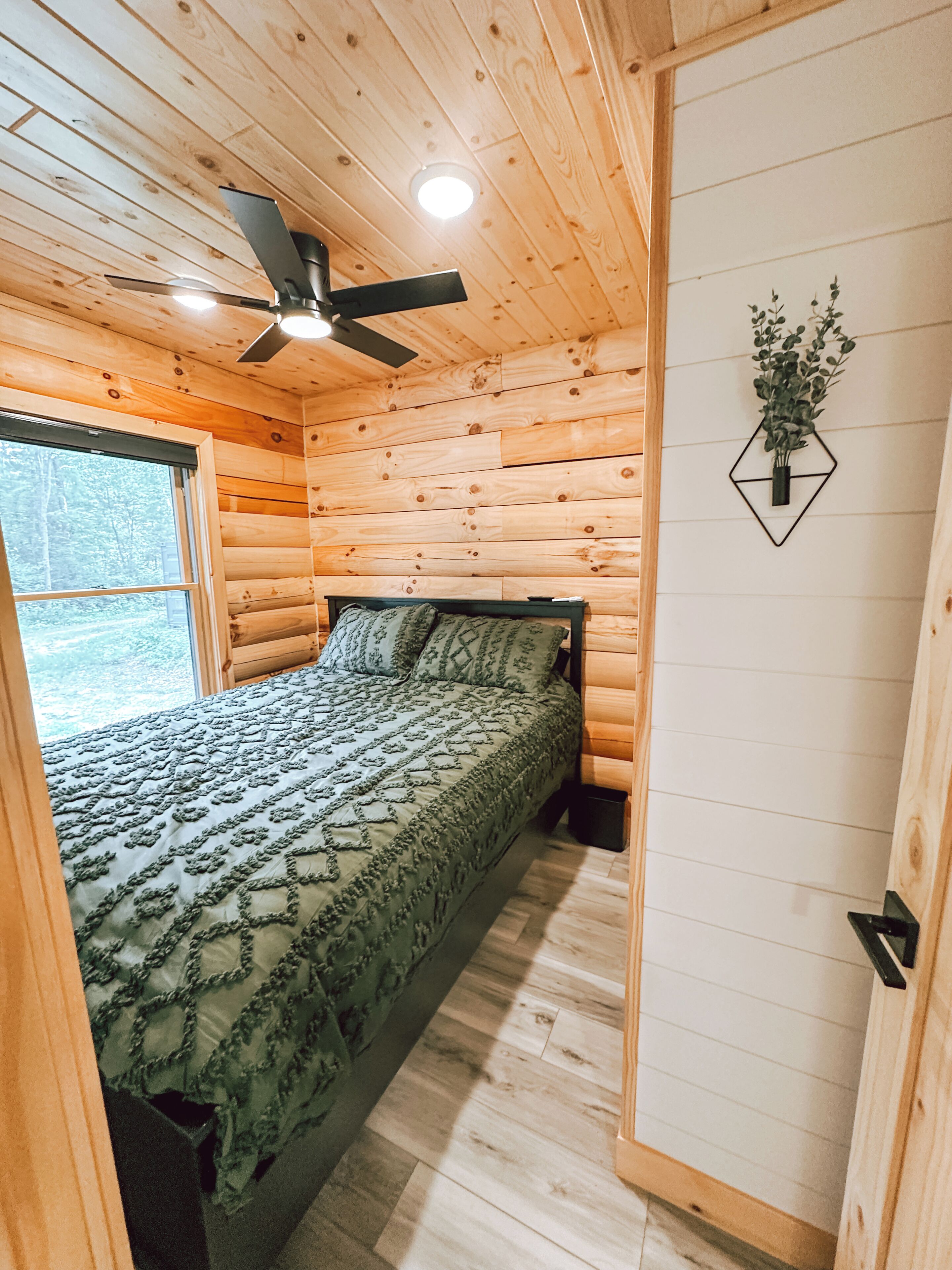 Quarto de cabana de madeira com uma cama, um ventilador de teto, uma janela e uma planta fixada na parede.