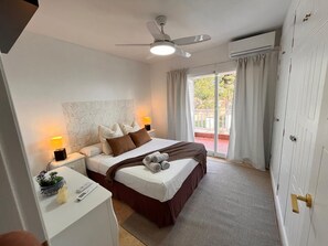 Ein Hotelzimmer mit Bett, Nachttischlampen, Deckenventilator, Klimaanlage, einem kleinen Schreibtisch mit Pflanze und Blick ins Freie.