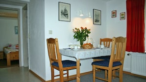 Un comedor con una mesa lista para dos comensales, dos sillas de madera con cojines azules, un jarrón con flores y una lámpara fijada a la pared.