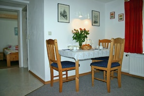 A dining area with a table set for two, two wooden chairs with blue cushions, a vase with flowers, and a wall-mounted light fixture. - Living under a thatched roof, in the quiet neighborhood of Fahretoft, 8 km from the North Sea (Dagebüll)