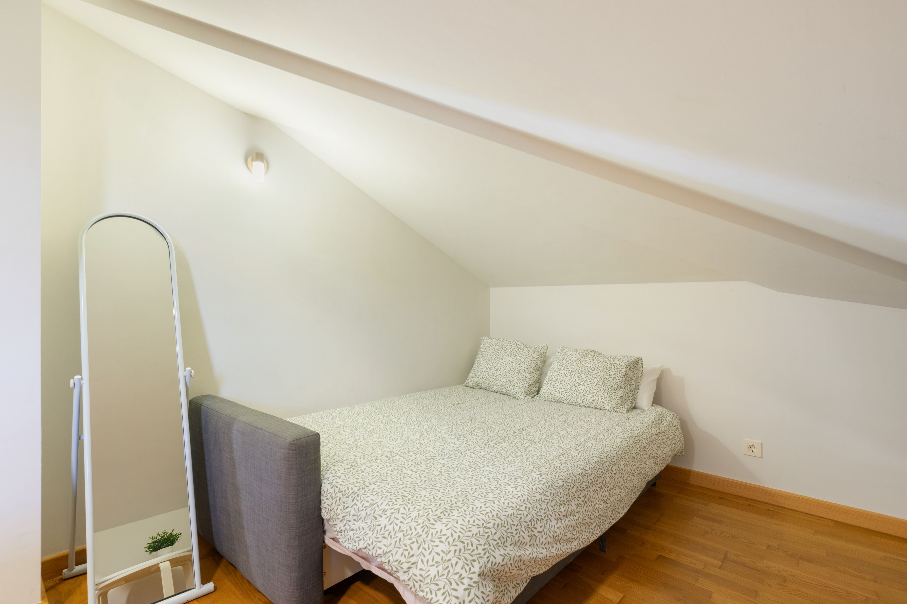 A small, single-bed room with a sloped ceiling, a standing mirror, and a grey bench.