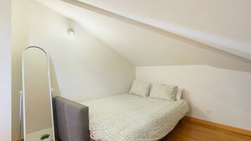 A small, single-bed room with a sloped ceiling, a standing mirror, and a grey bench.