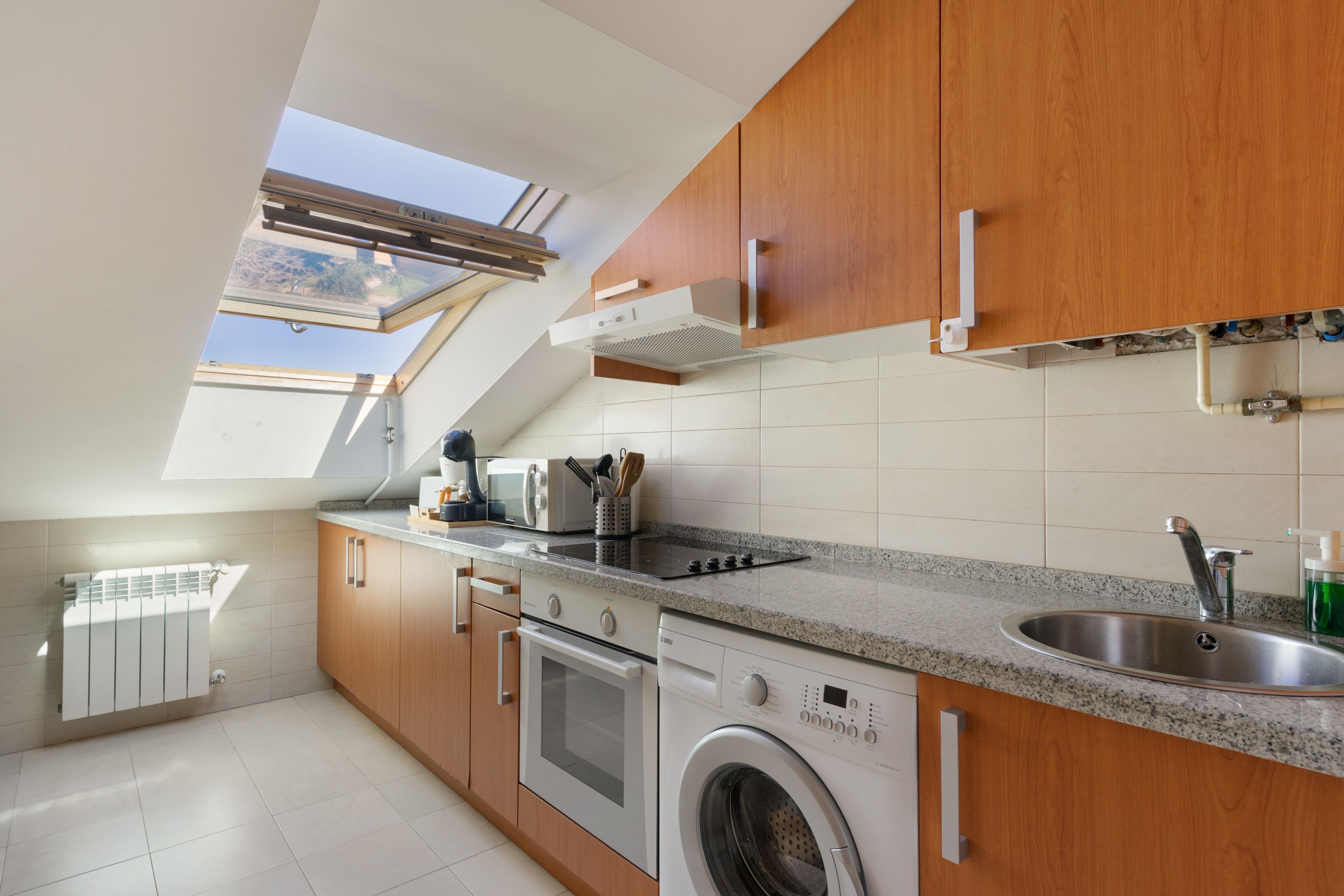 A modern kitchen with wooden cabinets, a built-in oven, a washing machine, and a skylight.