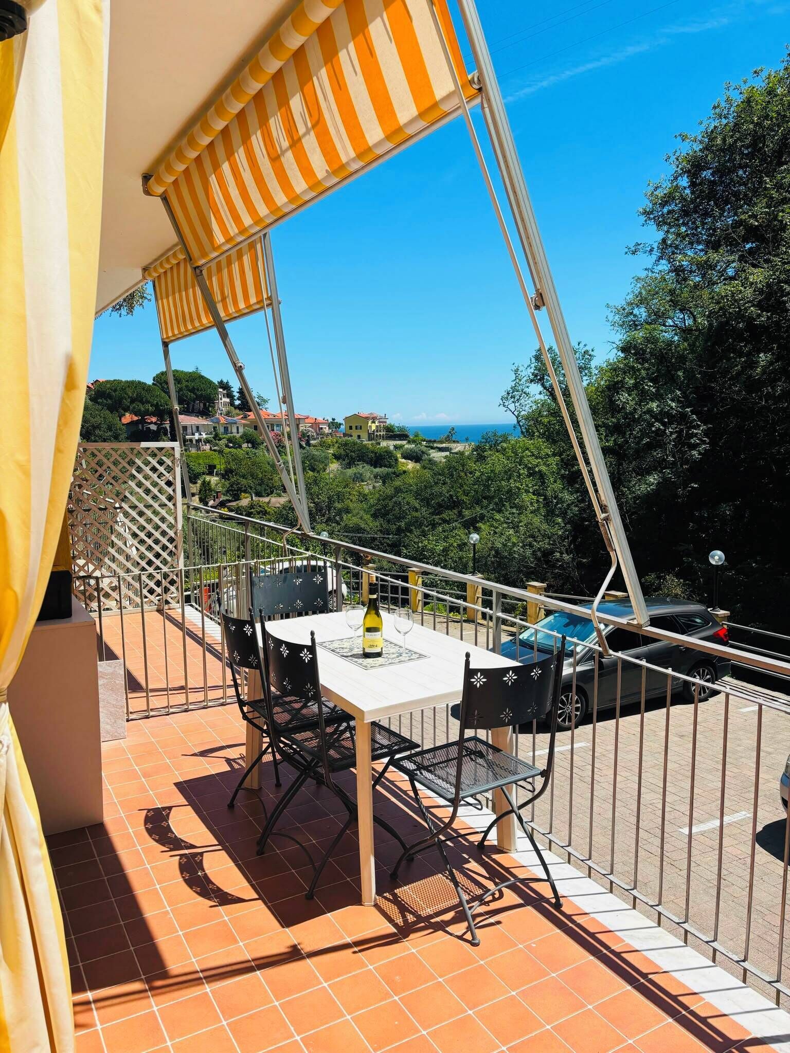 Una terrazza con tavolo, sedie e una bottiglia di vino, affacciata su una zona costiera con edifici e alberi.
