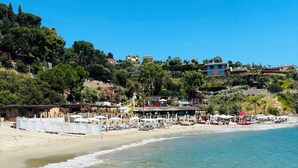 Ein Strand mit klarem Wasser, Sonnenschirmen und Gebäuden im Hintergrund.
