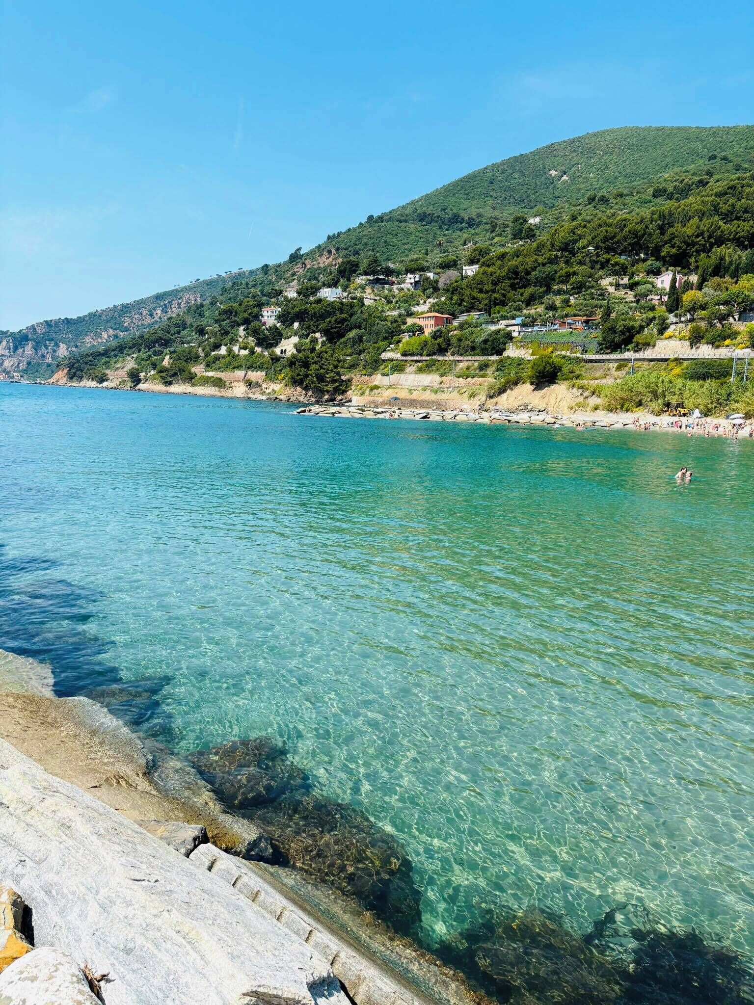 Eine Küstenlandschaft mit klarem Wasser, einer felsigen Küste und einem Hügel mit Gebäuden.