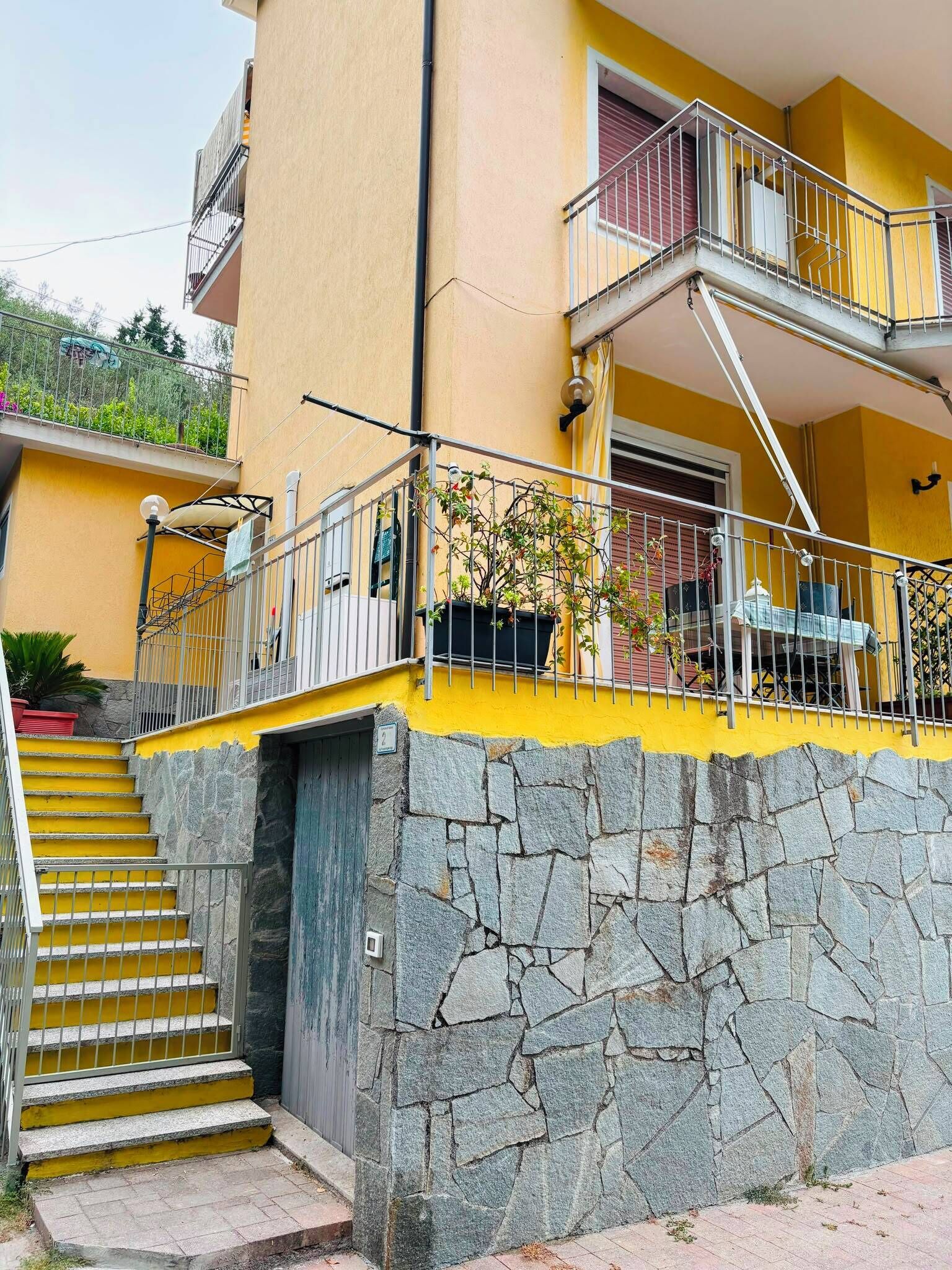 Un bâtiment à deux étages, avec une façade jaune, une base en pierre, des garde-corps en métal et un escalier menant à l’entrée.