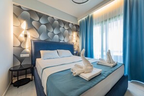 A modern hotel room with a large bed, a round side table, a wall-mounted light fixture, and a view of the city through sheer curtains.
