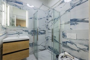 A bathroom with a glass shower enclosure, a marble-tiled wall, a wooden vanity with a sink, and a hairdryer on a small table.