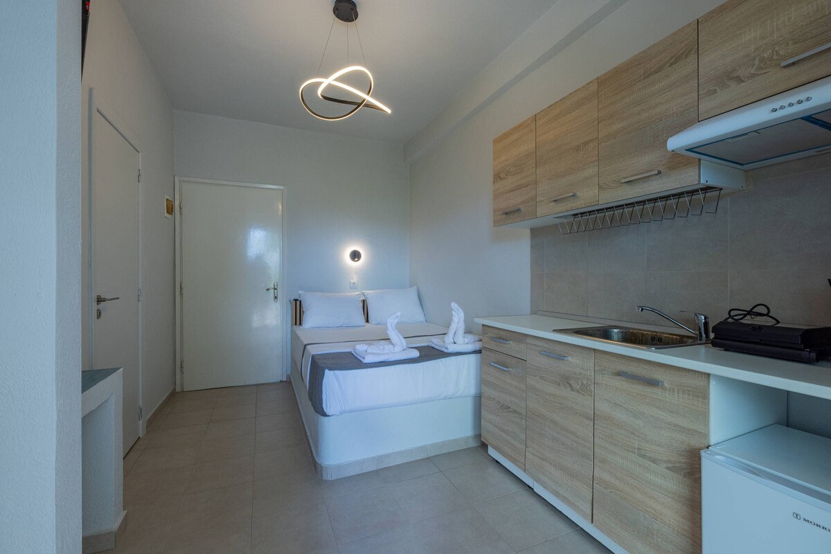 A compact hotel room with a bed, a kitchenette, and a modern pendant light.