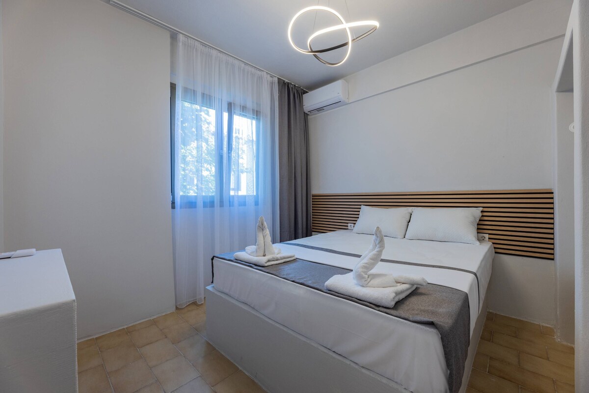 A hotel room with a bed, a bedside table, a window with curtains, and a modern light fixture.
