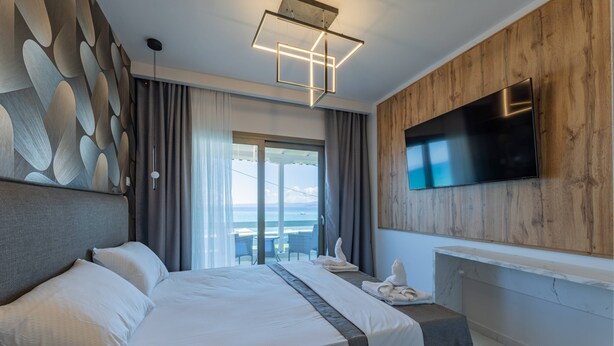 A modern hotel room with a large bed, a flat-screen TV mounted on the wall, and a view of the sea through the window.