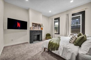 Une chambre à coucher avec un téléviseur fixé au mur, une cheminée, un lit avec des oreillers et une fenêtre avec des rideaux.