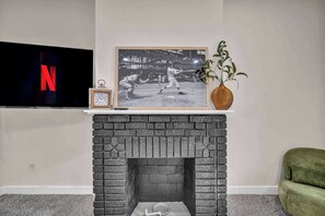A living room with a fireplace, a framed baseball photo, a clock, a vase with a plant, and a TV displaying the Netflix logo. - Near Tremont - 4BR - Sleeps 15 - Xbox (Cleveland)