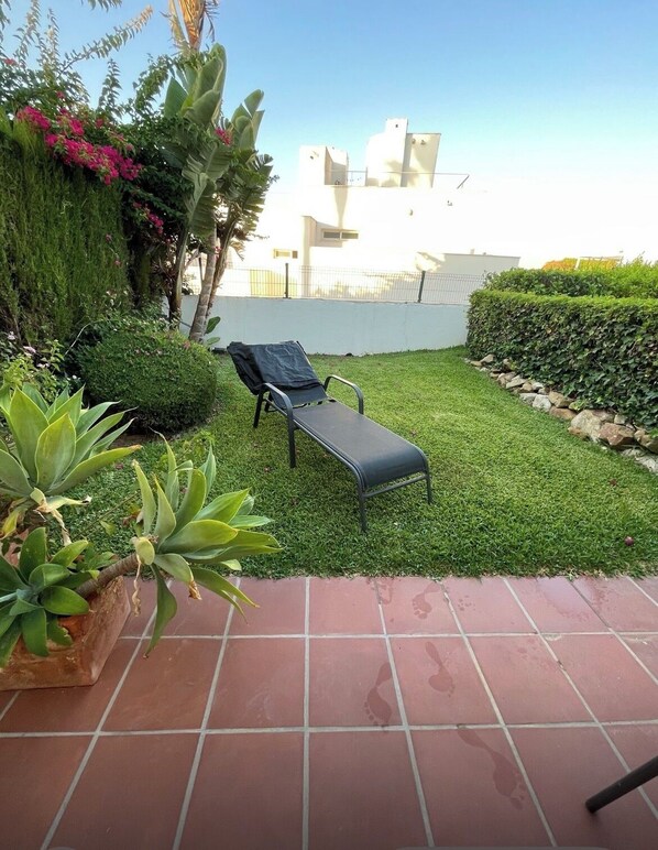 Eine Terrasse mit Liegestuhl, ein kleiner Garten mit verschiedenen Pflanzen und ein gefliester Boden.